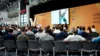 Vortrag auf der IAA. Menschen sitzen in Reihen in einer Halle. Vorne ist eine Leinwand.