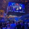 Das Bild zeigt eine große Menschenmenge in einer Arena bei einem Live-Event. Im Mittelpunkt des Bildes hängt ein großer Bildschirm von der Decke, auf dem mehrere Personen auf einer Bühne zu sehen sind. Die Arena ist voll besetzt, das Publikum beobachtet gespannt die Bühne und den Bildschirm. Die Beleuchtung ist blau und es herrscht eine lebhafte Atmosphäre, die an ein eSport-Turnier oder eine große Gaming-Veranstaltung erinnert.