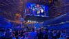 Das Bild zeigt eine große Menschenmenge in einer Arena bei einem Live-Event. Im Mittelpunkt des Bildes hängt ein großer Bildschirm von der Decke, auf dem mehrere Personen auf einer Bühne zu sehen sind. Die Arena ist voll besetzt, das Publikum beobachtet gespannt die Bühne und den Bildschirm. Die Beleuchtung ist blau und es herrscht eine lebhafte Atmosphäre, die an ein eSport-Turnier oder eine große Gaming-Veranstaltung erinnert.