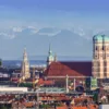Die Stadt München mit allen bekannten Sehenswürdigkeiten ist im Vordergrund abgebildet. Im Hintergrund sieht man die Alpen.