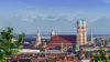 Die Stadt München mit allen bekannten Sehenswürdigkeiten ist im Vordergrund abgebildet. Im Hintergrund sieht man die Alpen.
